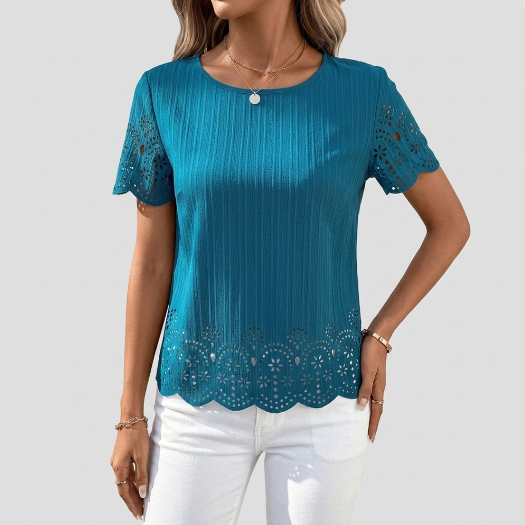 A woman wearing a teal blue short-sleeve top with vertical pleats, scalloped eyelet lace trim at the sleeves and hem, paired with white pants, against a light gray background.