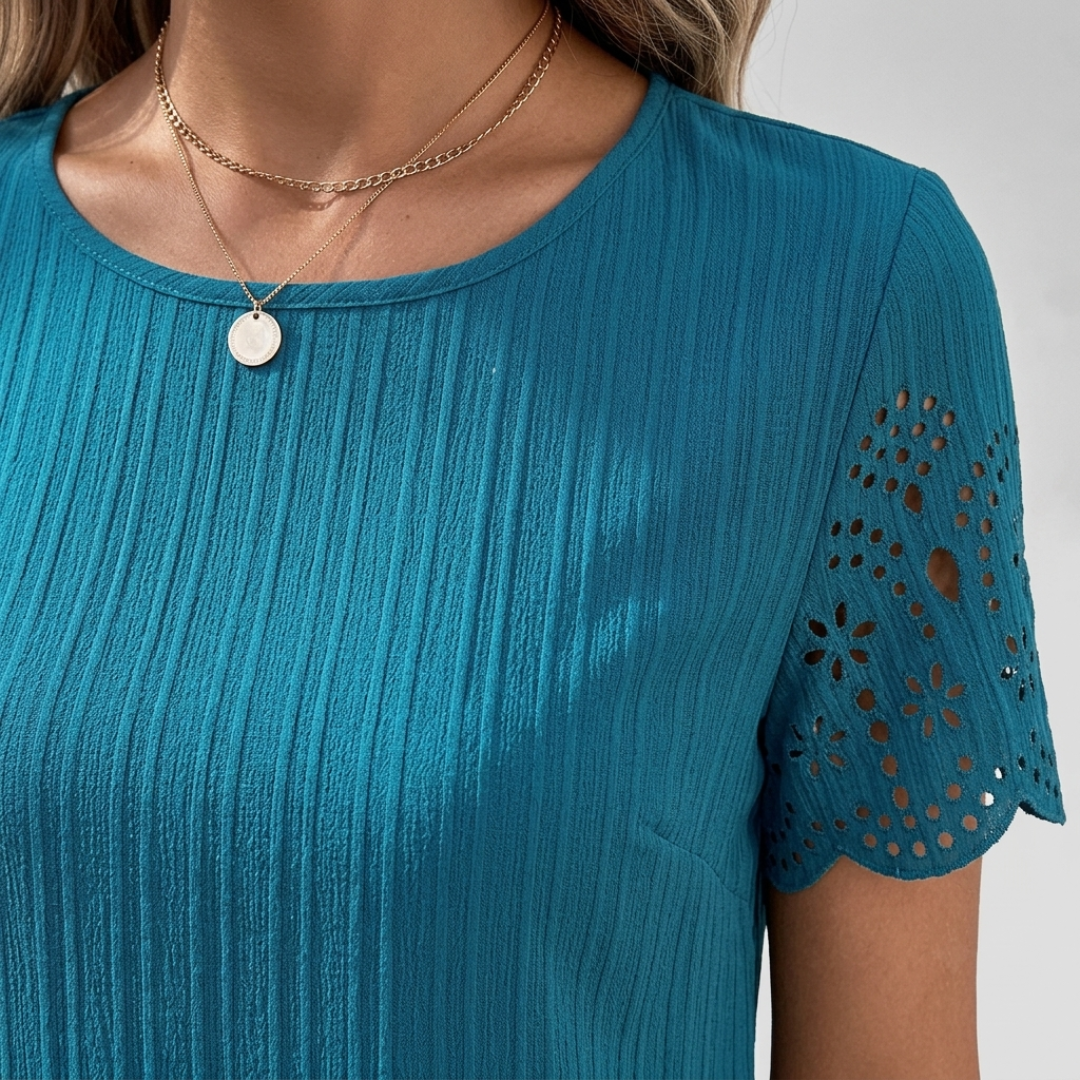 A close-up of a woman wearing a teal short-sleeve top with vertical pleats and floral eyelet lace sleeves, styled with layered gold necklaces.
