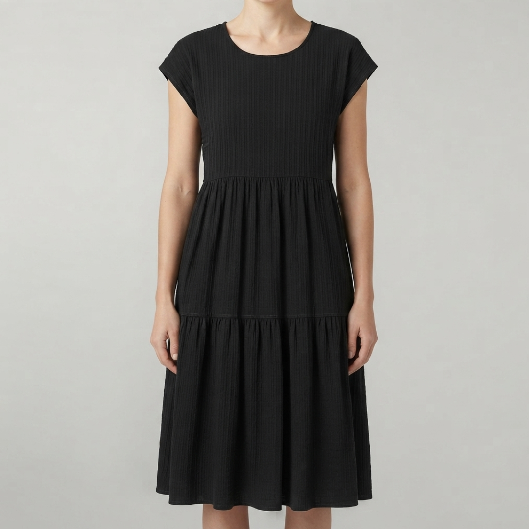 A woman wearing a black tiered dress with cap sleeves and a gathered waist, photographed against a plain light gray studio background.