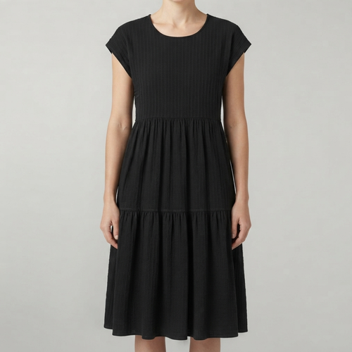 A woman wearing a black tiered dress with cap sleeves and a gathered waist, photographed against a plain light gray studio background.
