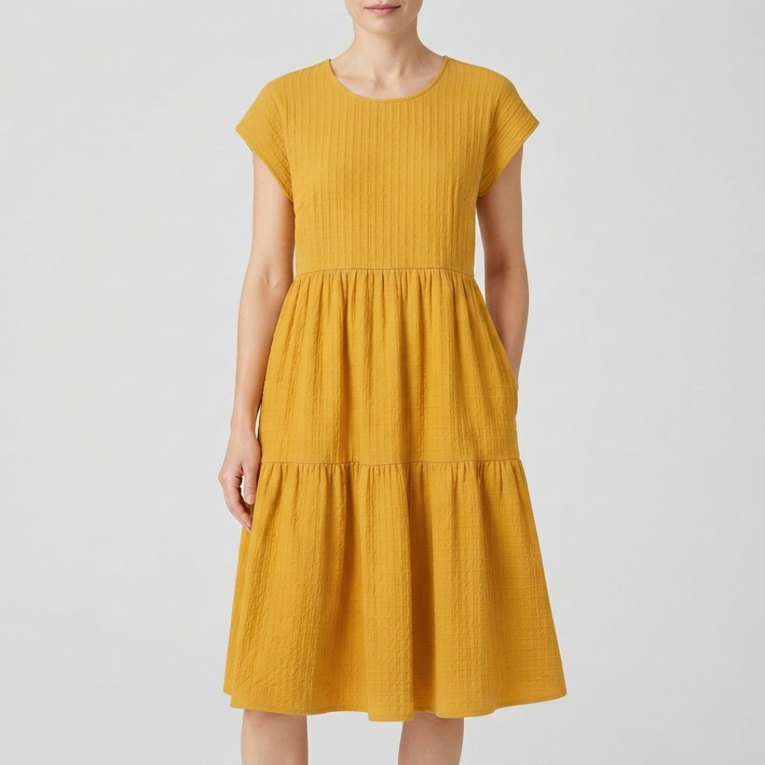 A woman wearing a mustard yellow textured tiered dress with short cap sleeves and a round neckline, photographed against a light gray studio background.