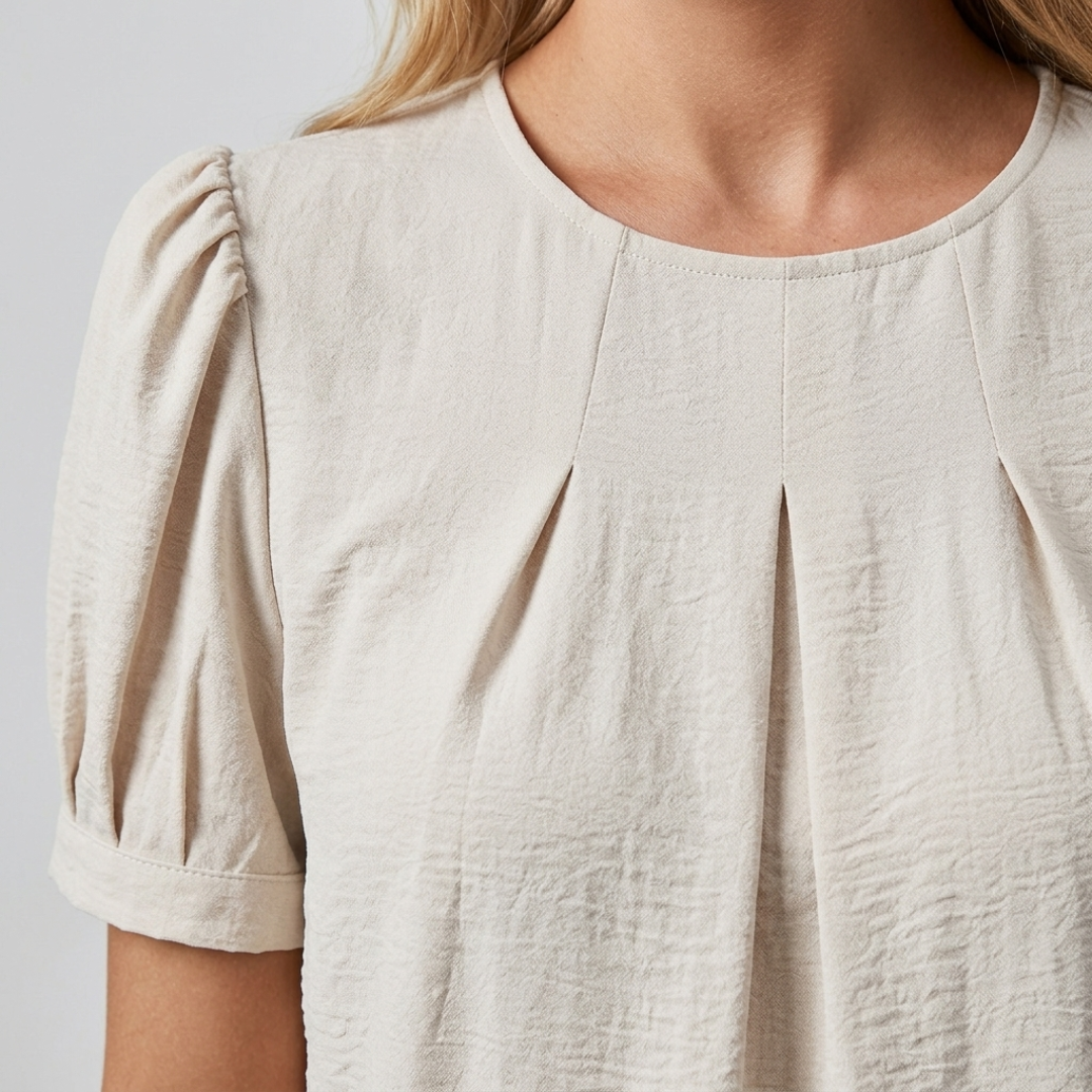 Close-up of a woman wearing a light beige short-sleeve blouse with gathered puff sleeves and a round neckline, shown against a plain light gray background.