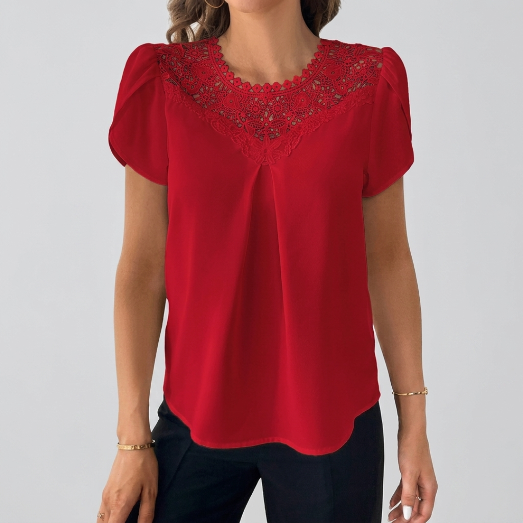 A woman wearing a bright red short-sleeve blouse with a scalloped lace V-neckline detail, paired with dark pants, against a plain light gray studio background.