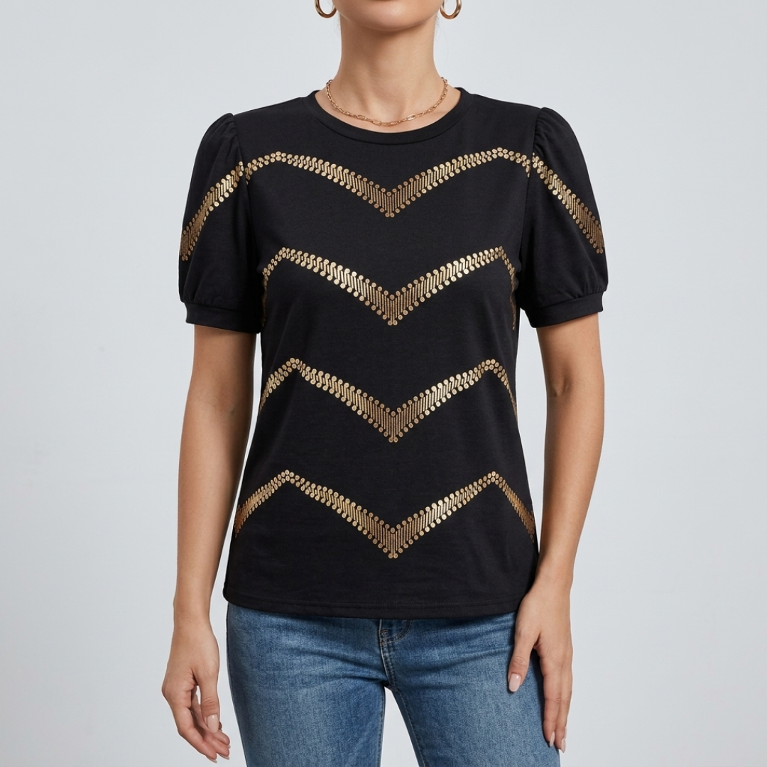 A woman wearing a black short-sleeve top with metallic gold chevron patterns across the front, paired with blue jeans, against a light gray studio background.