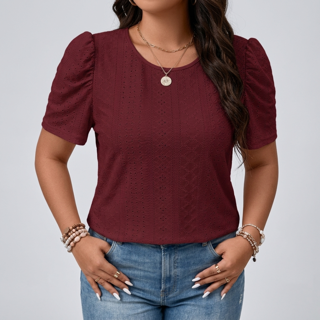 A woman wearing a short-sleeve burgundy eyelet knit top with puff sleeves, paired with blue jeans and layered gold necklaces, against a light gray studio background.