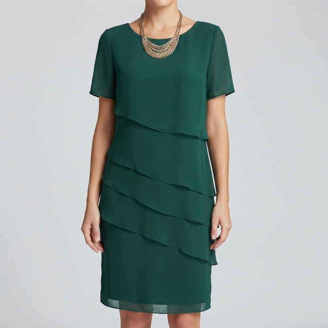 A woman wearing a dark green short-sleeve shift dress with a layered ruffle skirt, styled with a multi-strand gold necklace, against a light gray studio background.
